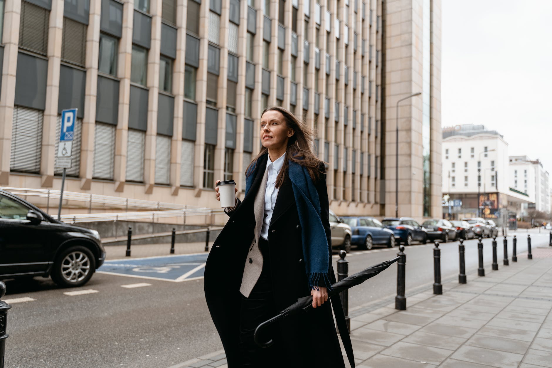 a woman in black blazer holding folded umbrella and cup of coffee while walking on the side of the road